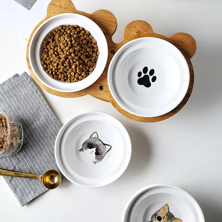 Set of ceramic pet bowls with playful designs, including a paw print and cat illustrations, placed on a wooden stand. Stylish food bowls for cats and dogs.